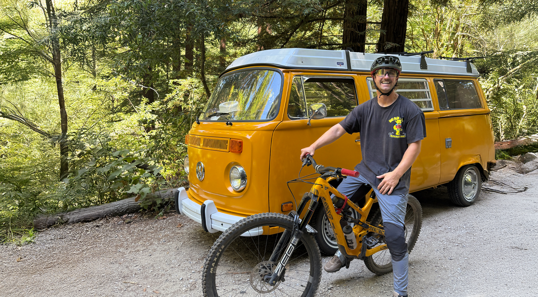  A man wearing a helmet and riding gear is standing next to the vanlife vintage Volkswagen van and a mountain bike on a dirt path in a wooded area.