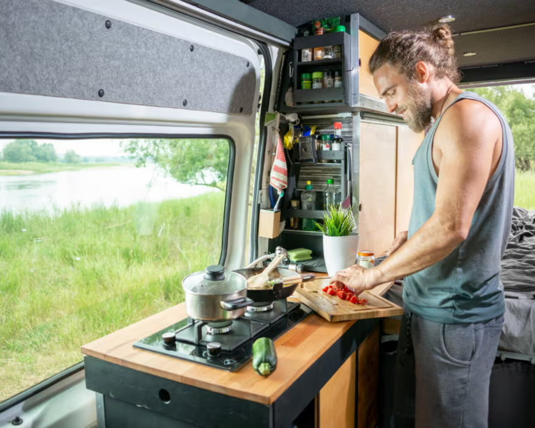  man cooking in the kitchen of his van with the window revealing grass in the background