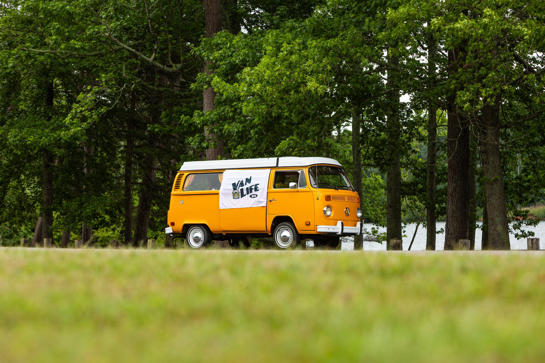 the vintage vanlife Volkswagen mango van with a white the vanlife logo on the side is parked on a low grassy hill near a body of water, backed by dense green trees.