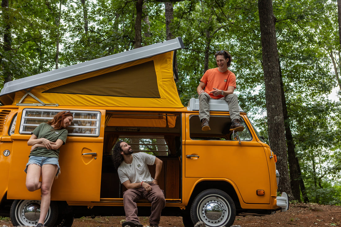 A woman, a man sitting on the roof, and another man sitting in the doorway of the vanlife mango Volkswagen camper van, all appearing relaxed in a forest setting.