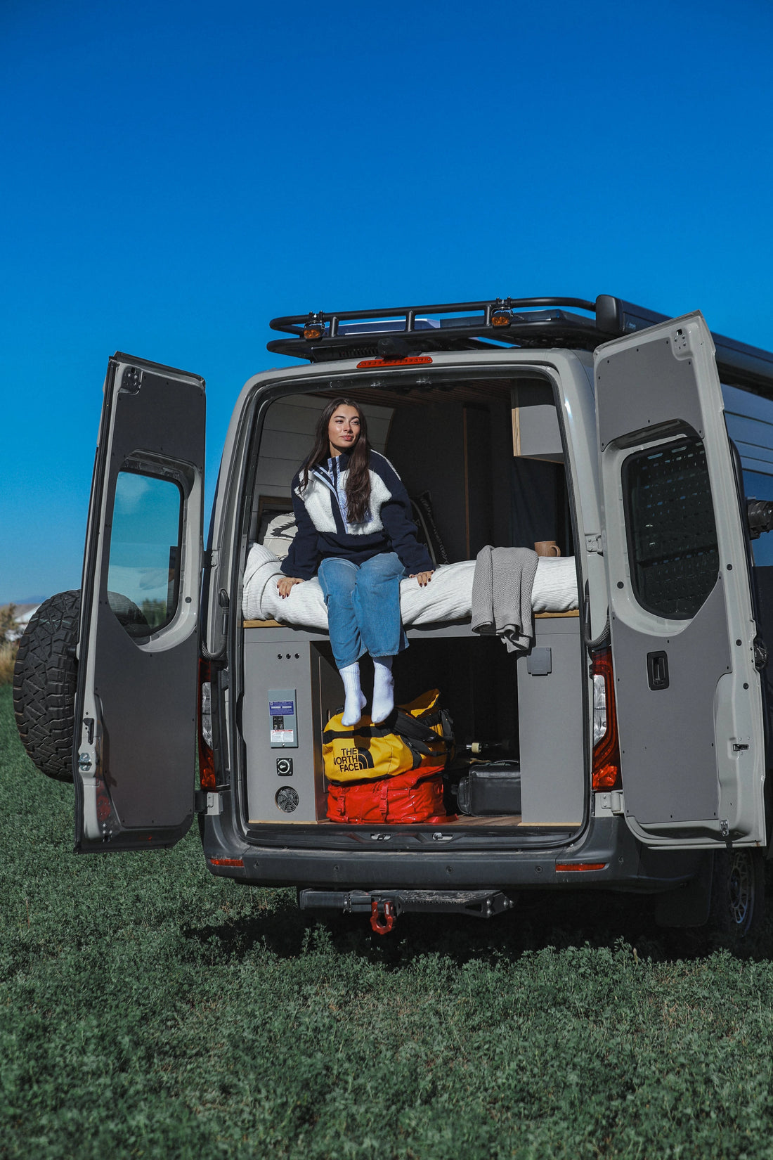 Woman sitting in the open door of a Sprinter van with camping gear inside, on grass with blue sky.