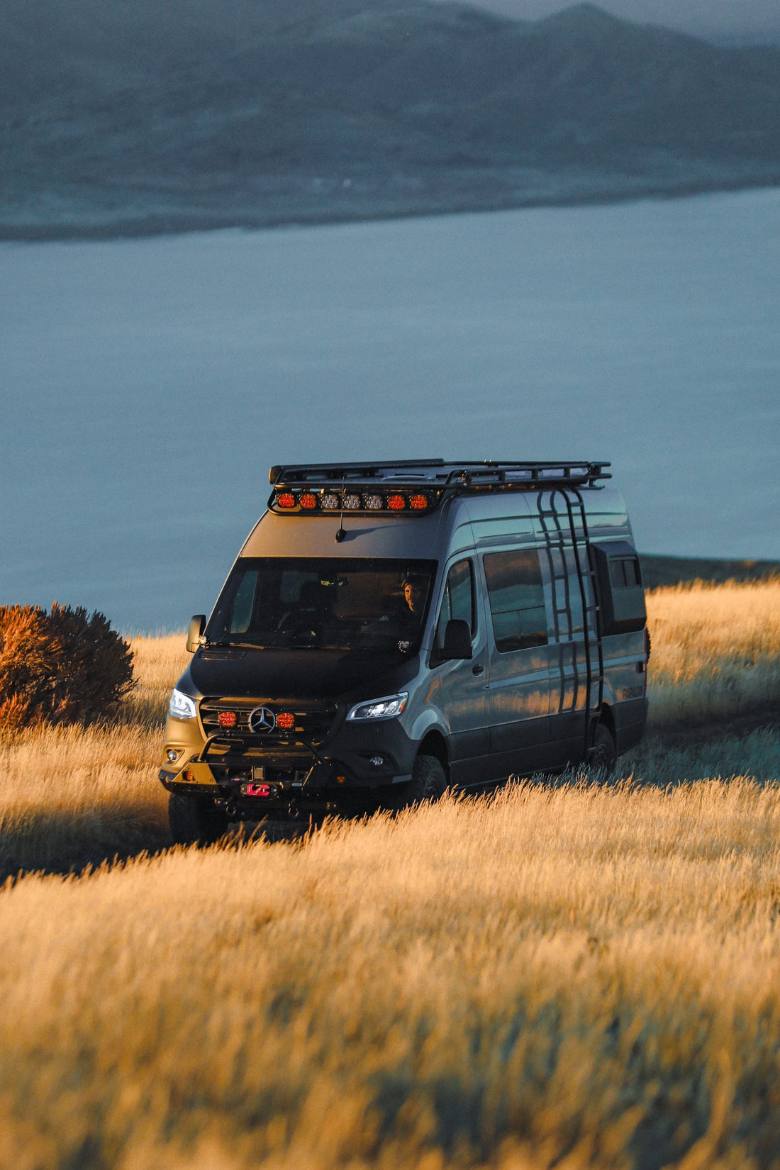 Mercedes Benz Sprinter van with roof rack in a grassy field with mountains and water in the background