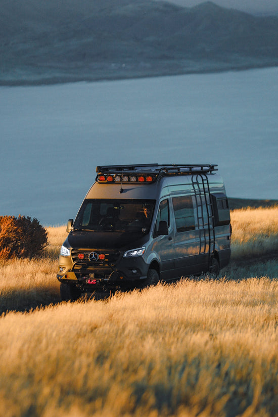 Mercedes Benz Sprinter van with roof rack in a grassy field with mountains and water in the background