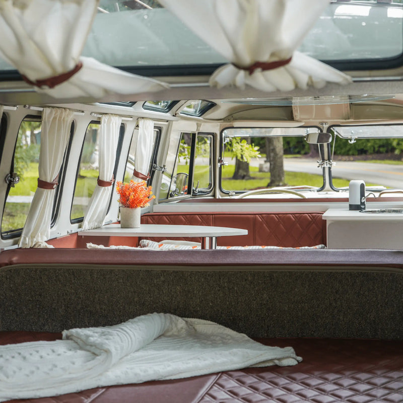 Looking forward through the prize Bus from the rear bench: a folded blanket, a table with orange flowers and curtains tied back at the windows