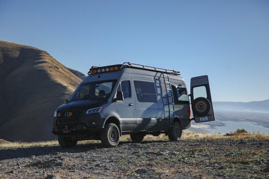 Mercedes Benz Sprinter van with open doors on a scenic landscape with mountains and a lake in the background.