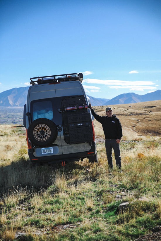 Man standing next to a Mercedes Benz Sprinter van in a grassy field with mountains in the background